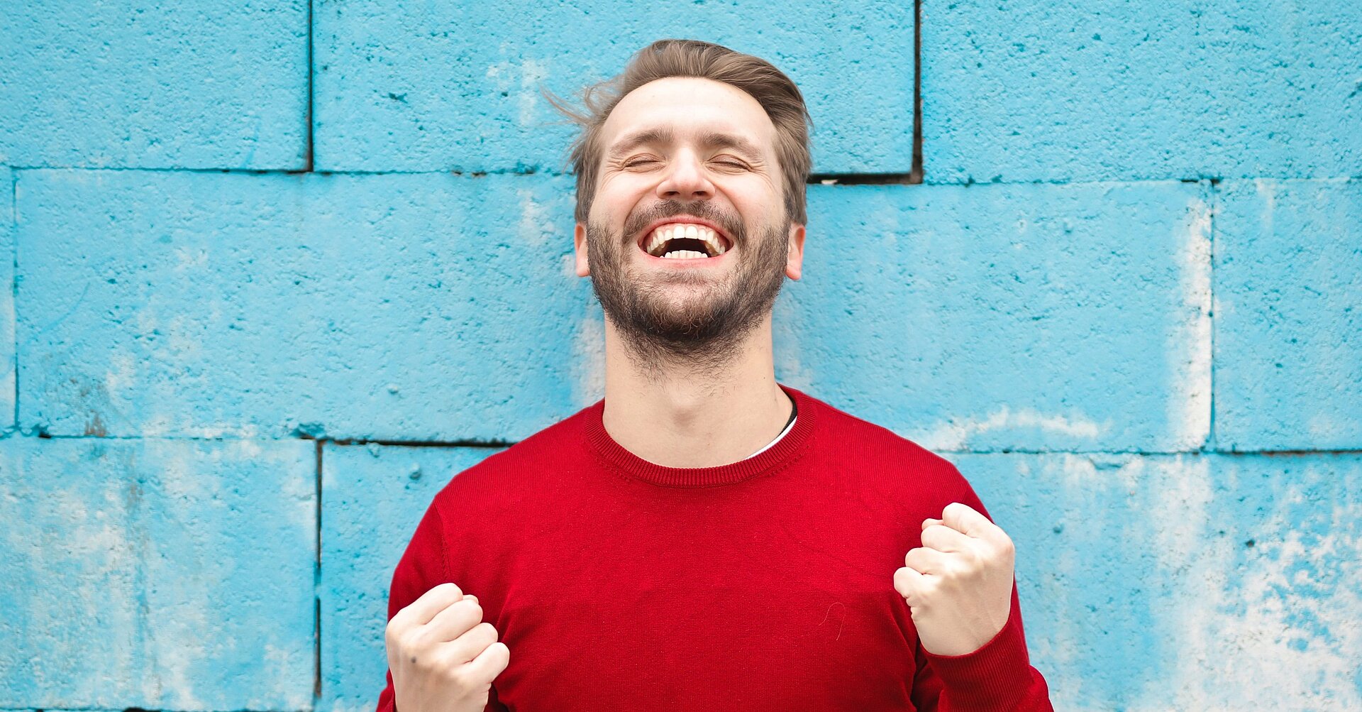 	Foto eines Mannes in einem roten Pullover vor einer blauen Mauer, der die Hände zu Fäusten ballt und lacht.